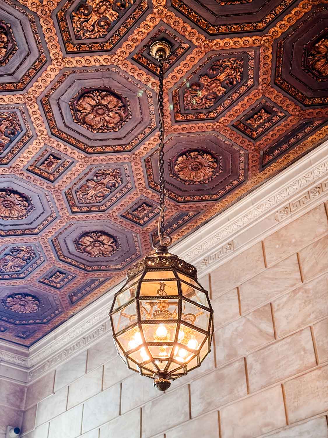 A large glass chandelier hanging from a ceiling of ornately carved wood, sits above a stairwell in the NYPL.