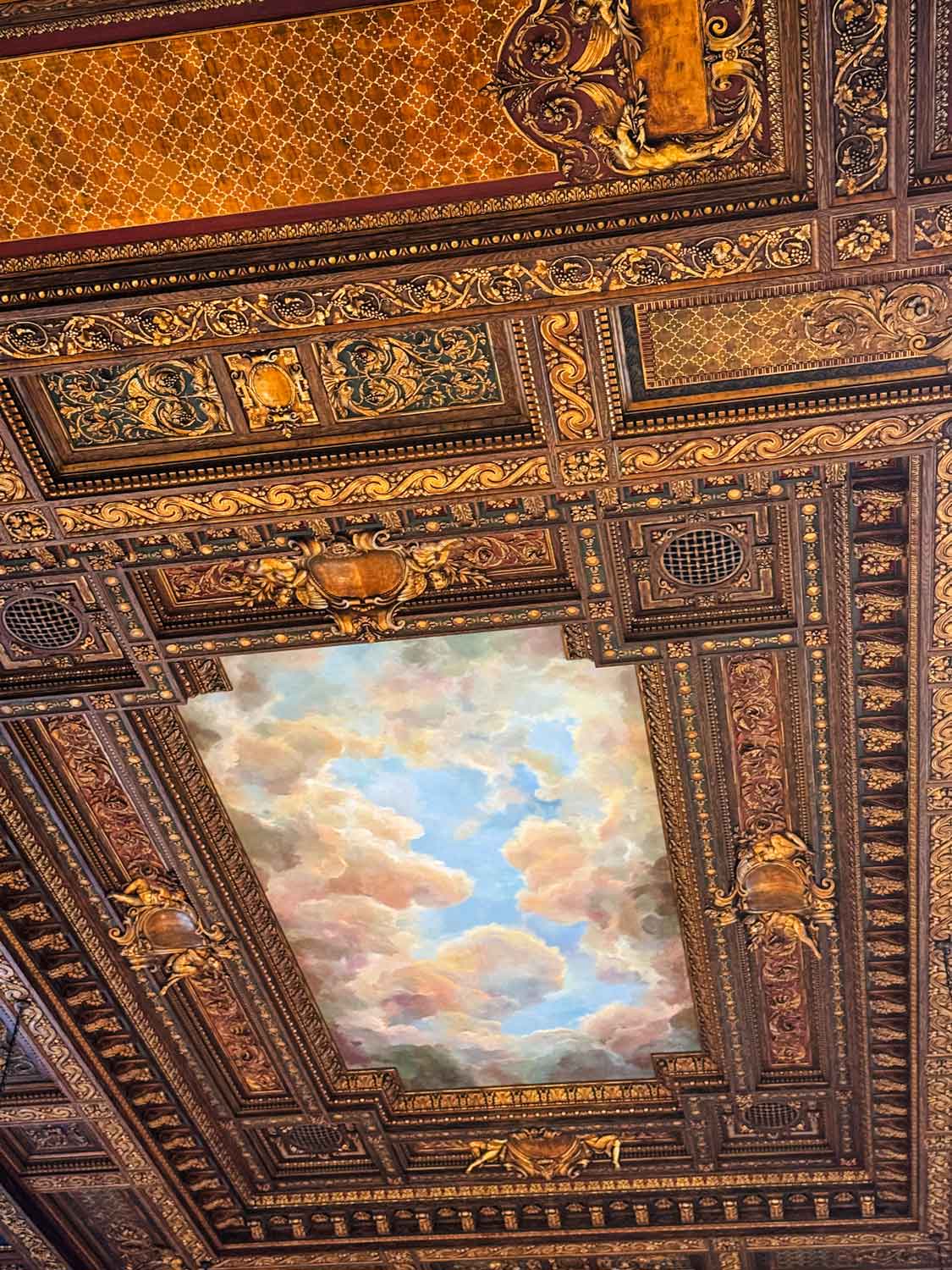 The ceiling of the rose reading room has a mural of blue sky and clouds. Ornamented carved wood with gold-plated frames the mural.