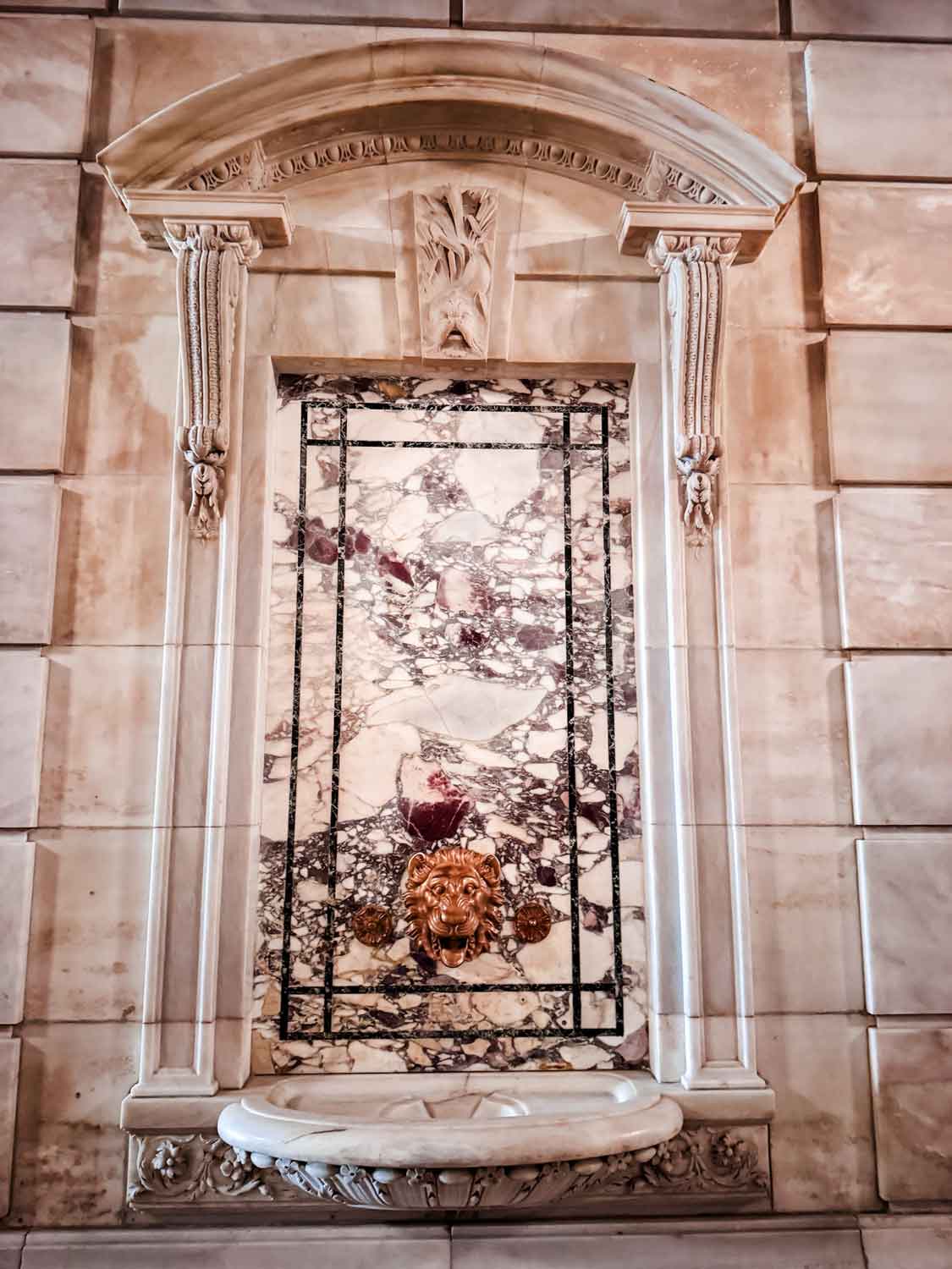 A marble water fountain with a gold Lion's head for the spout is on a granite wall at the NYPL.