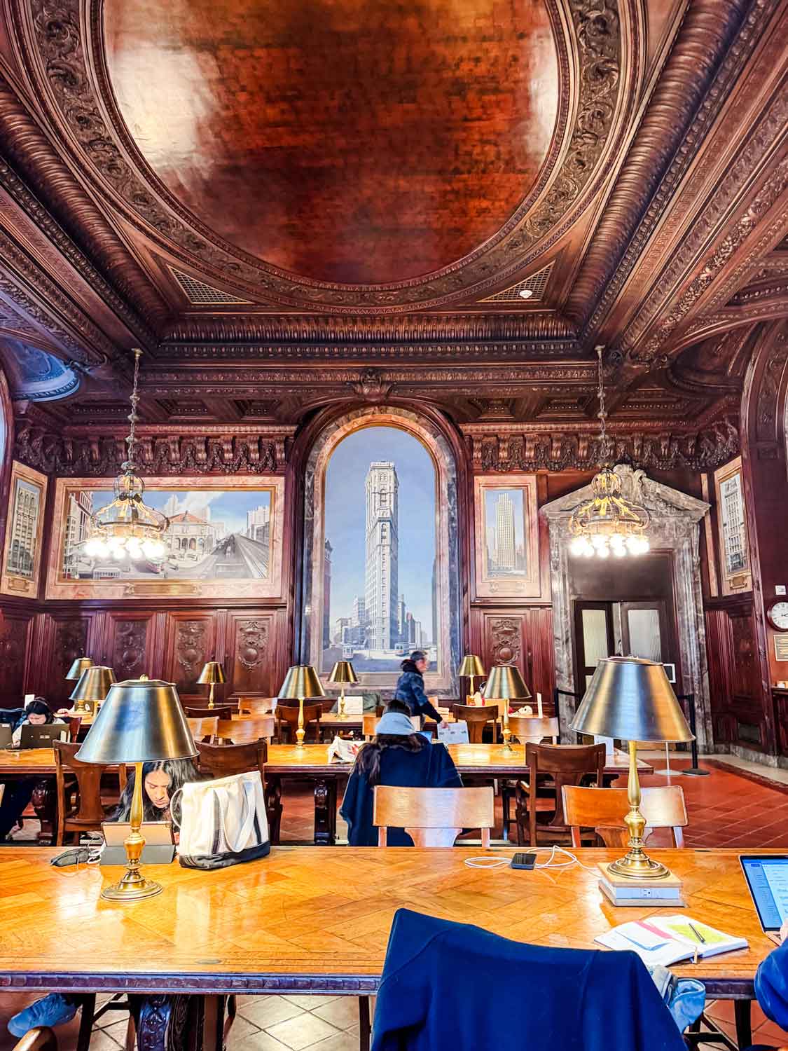 A reading and study room of the New York Public Library is filled with wooden tables with lamps on them and people working and studying.