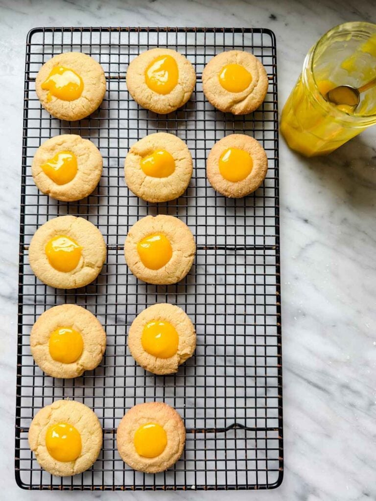 Thumbprint cookies have been baked and are on a wire cooling rack, and the indentation has been filled with homemade lemon curd.