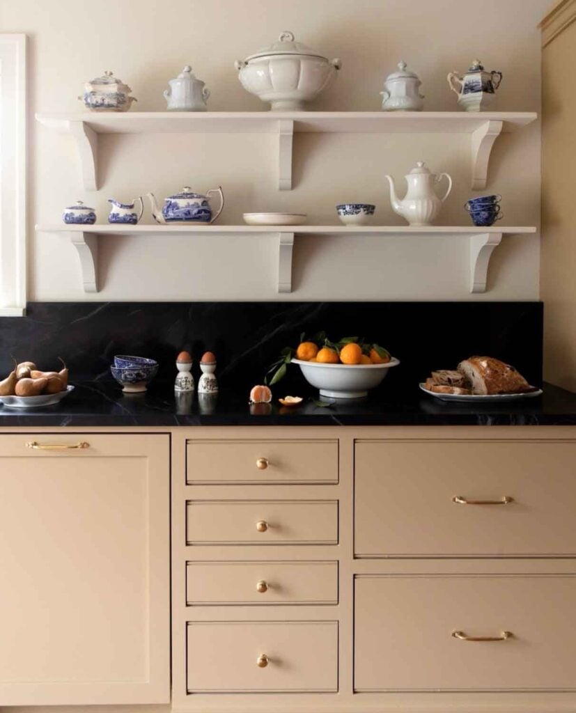 Open shelving full of tasteful pottery is above a counter with a bowl of fruit, a plate of pastries on top of it.