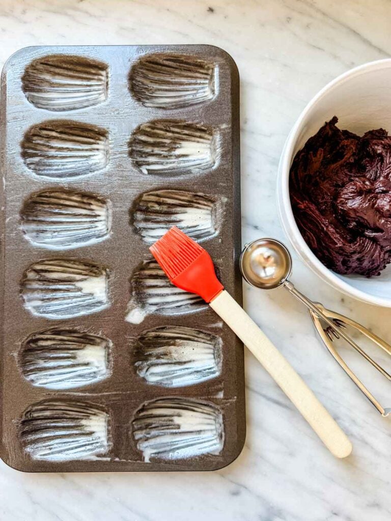 The shell molds in a Madeleine pan have been brushed with melted butter. A cookie scoop is on the counter next to a bowl of batter.