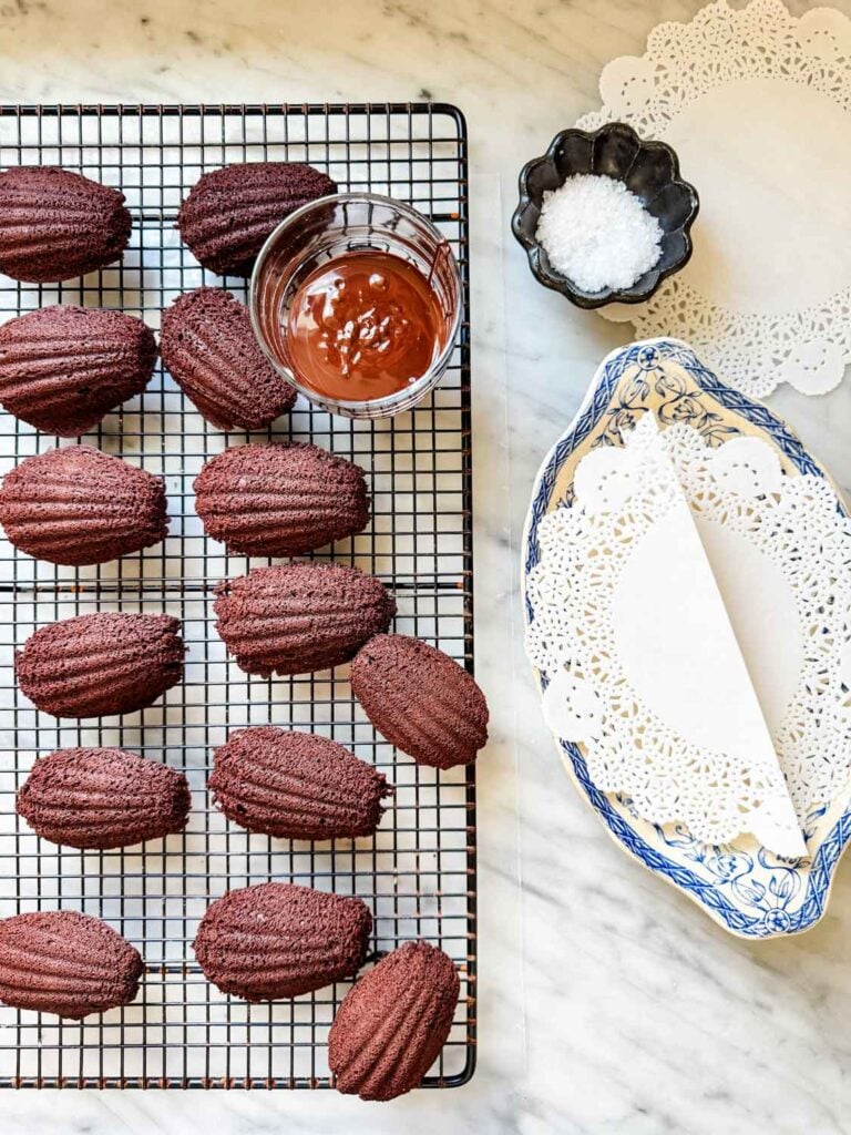 Madeleine cakes have been turned out on a wire cooling rack. On the rack is next to a glass with melted chocolate and is next to a bowl of sea salt, and a French vintage blue and white plate.