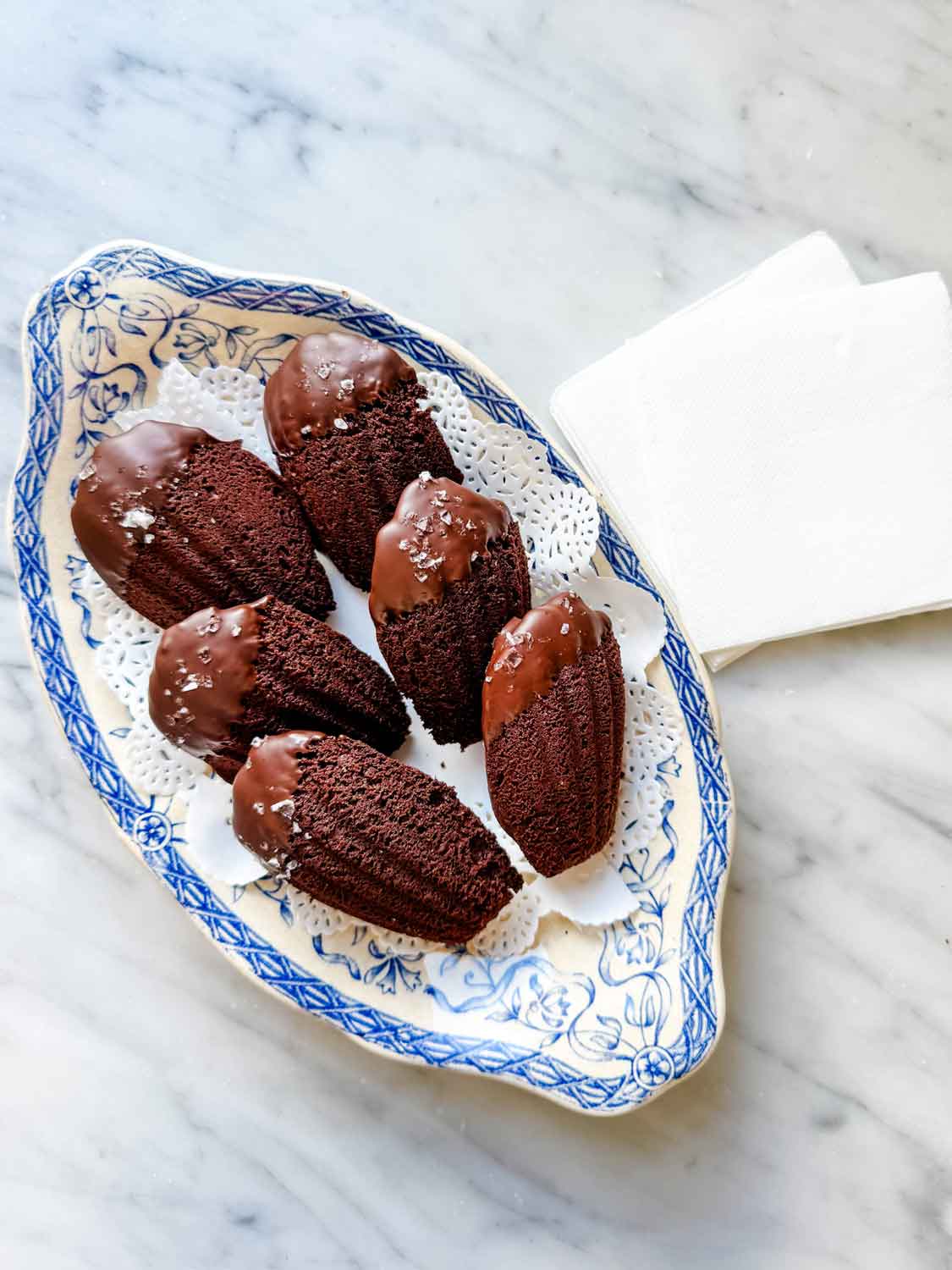 Six double dark chocolate madeleines, dipped in chocolate and sprinkled with sea salt, are served on a vintage French blue and white plate with a doily underneath the cakes.