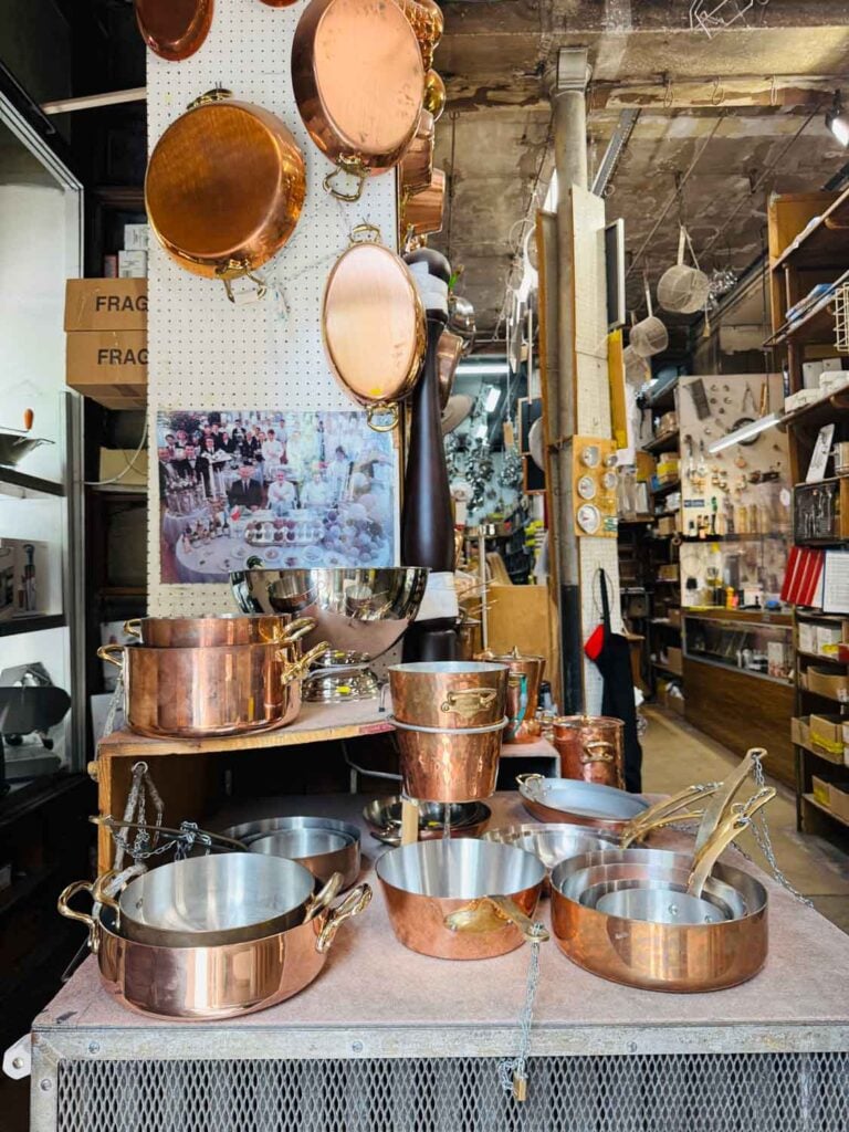 On top of a metal counter are stacks of different-sized and shaped copper cooking pans.