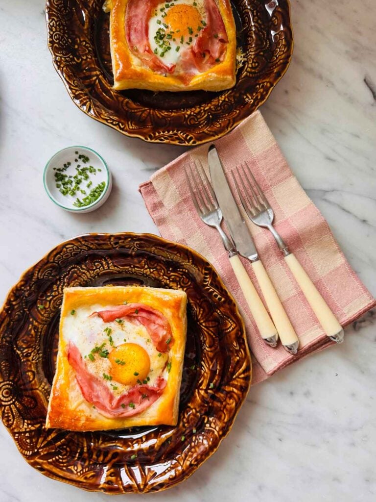 Puff pastry croque madame is topped with chives and served on small brown plates.
