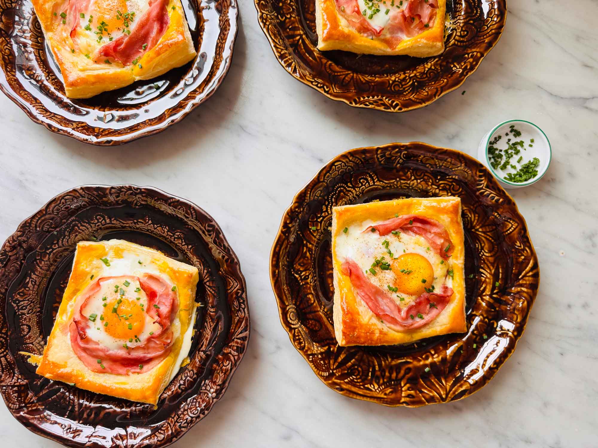 Puff pastry croque madame is topped with chives and served on small brown plates.