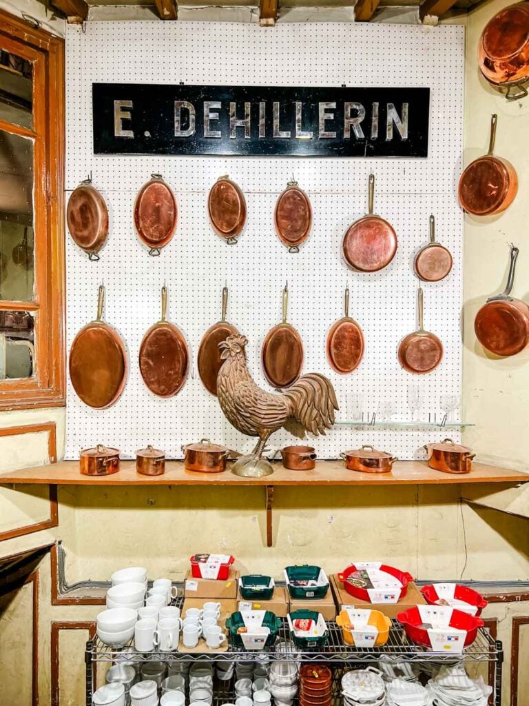 A metal E. Dehillerin placard hangs on a pegboard wall with various shapes and sizes of cooper pots and pans in this French kitchen supply store.