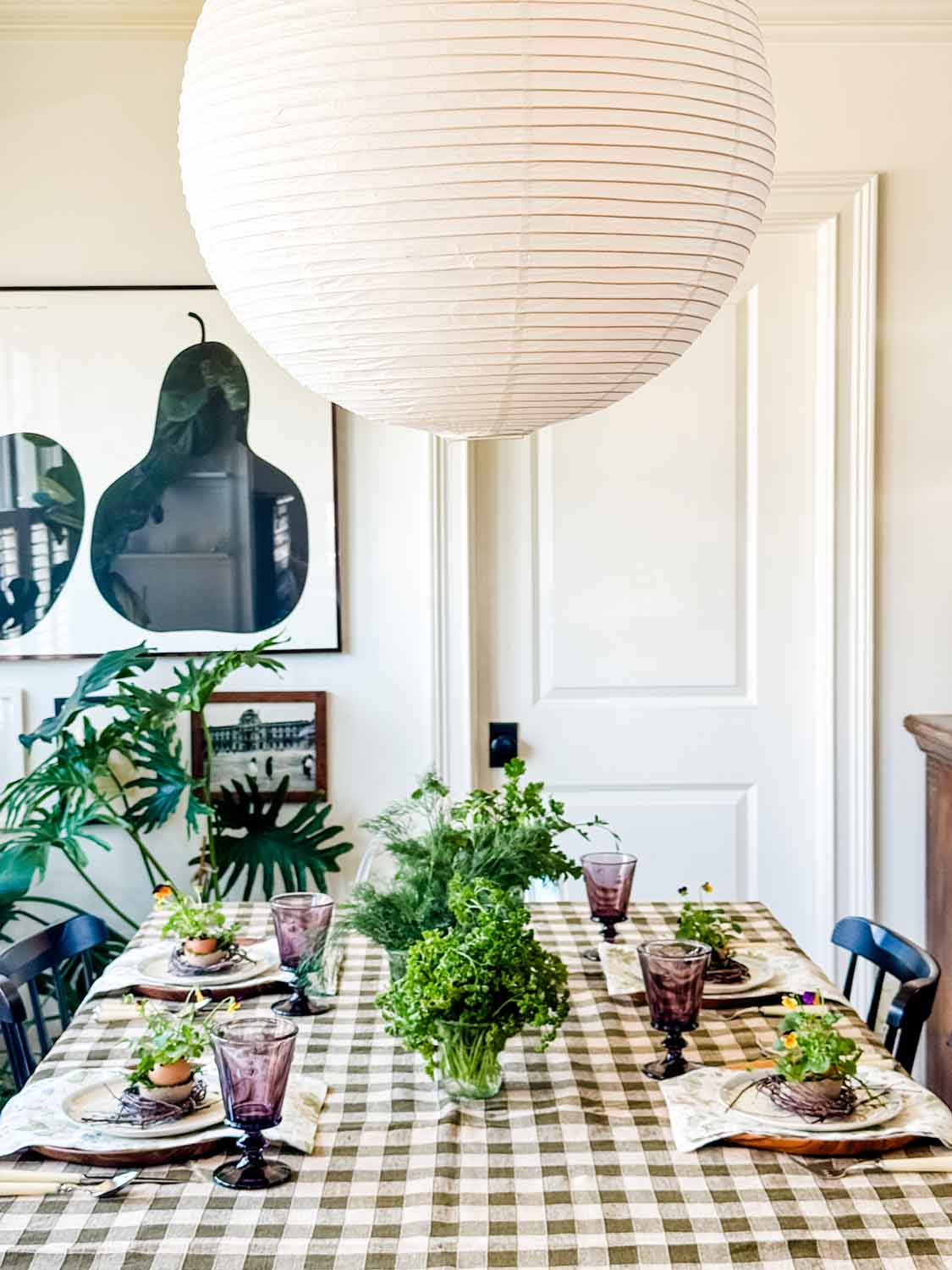 Dining room with globe paper pendant light, table with checked tablelcoth and black chairs. Table set with fresh herbs in jars.