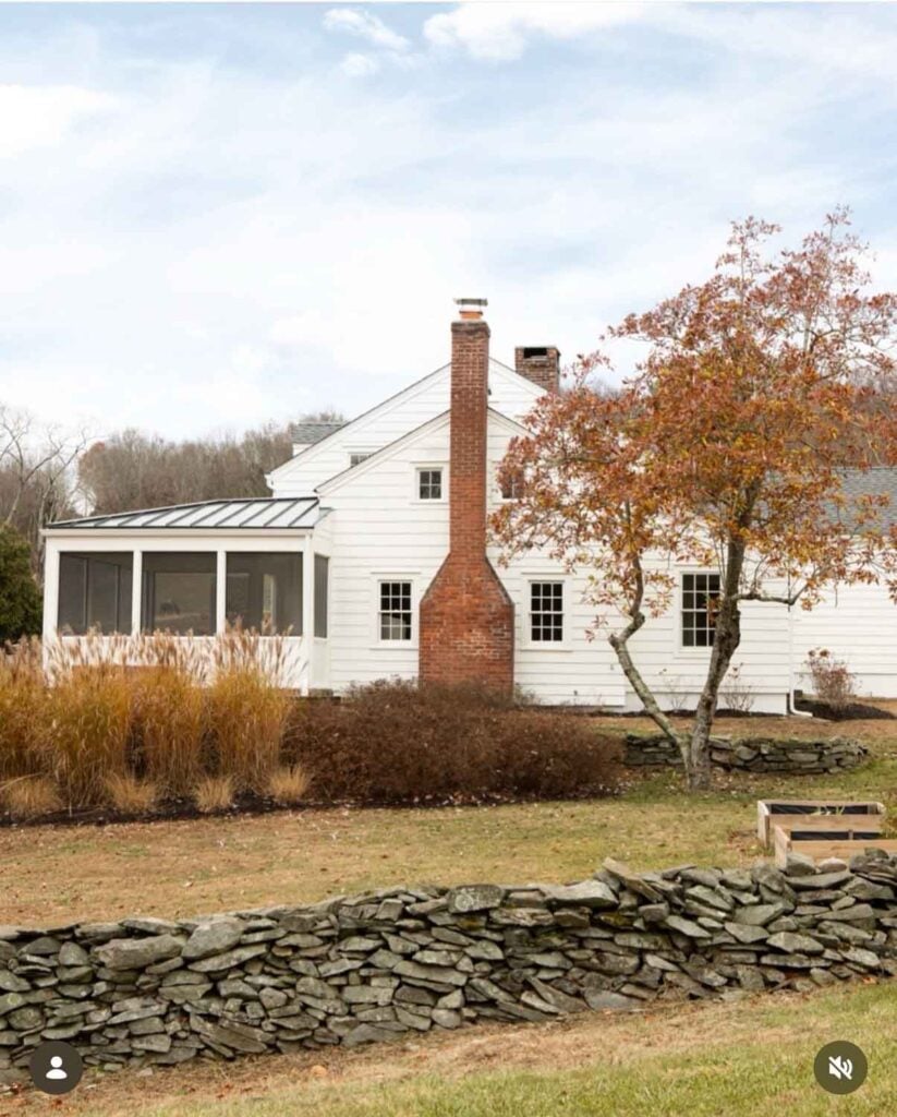 A rock wall runs along the exterior of this old white house in Hudson Valley, NY. A red brick chimney runs up the side of the house.