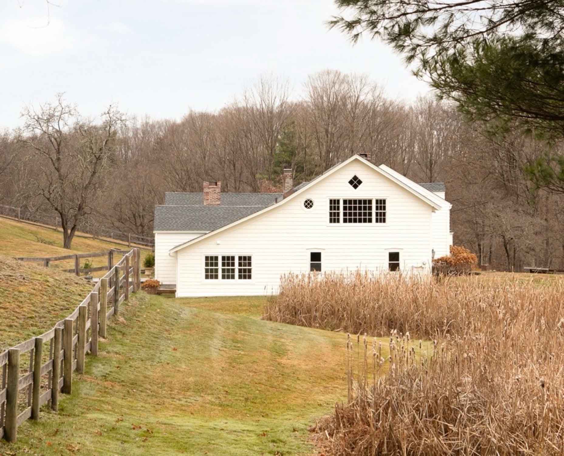 A white house in the Hudson Valley, NY, is surrounded by a fence, grass, and long grass. There are trees in the background.