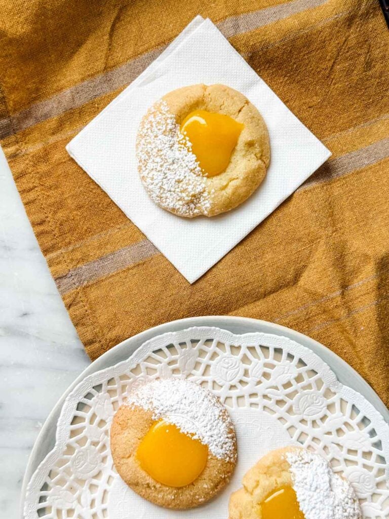 Lemon curd thumbprint cookies dusted with powdered sugar are sitting on a white napkin and a white plate with a round doily sheet.
