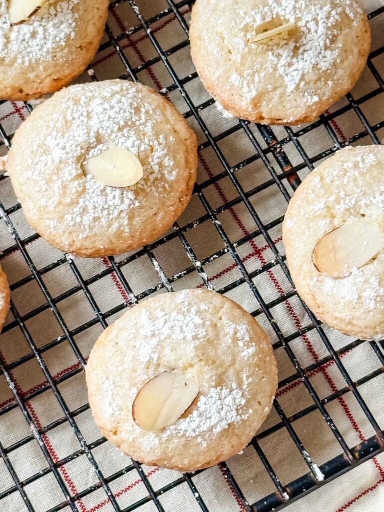 Macaron cookies with almond slivers are on a wire cooling rack that is sitting on top of a vintae French tea towel.