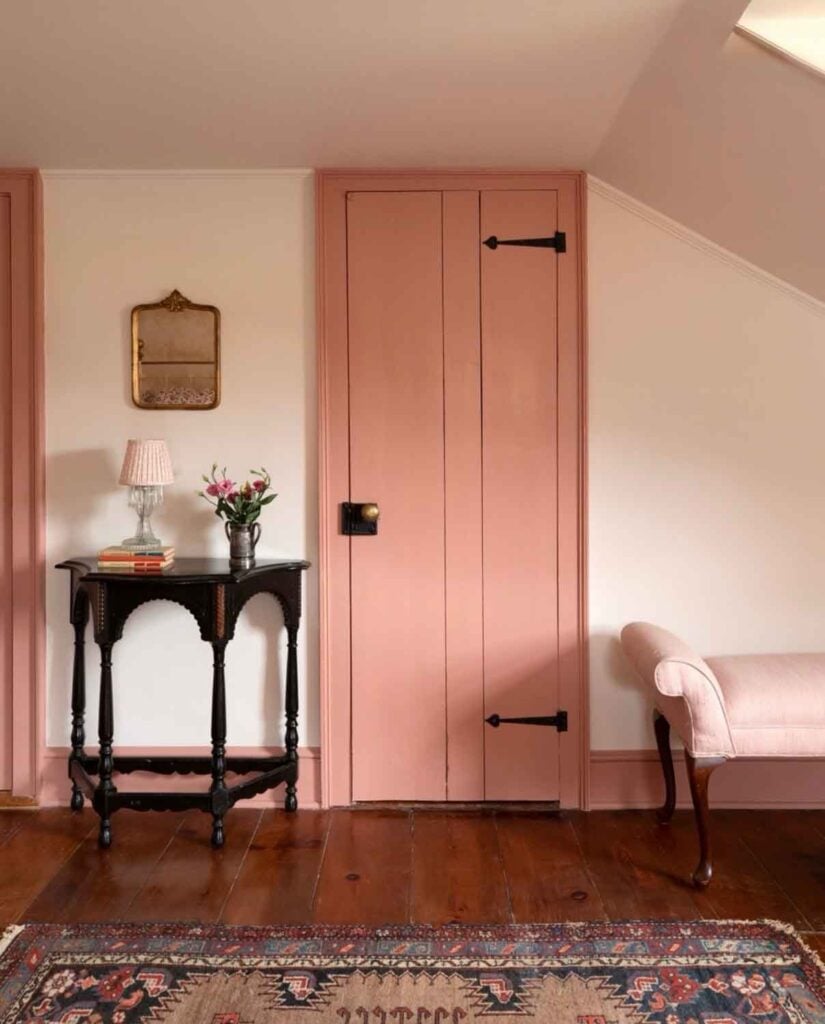 A corner table with a small lamp and a vase of floors is next to a old barn-stryle door with strap hinges.