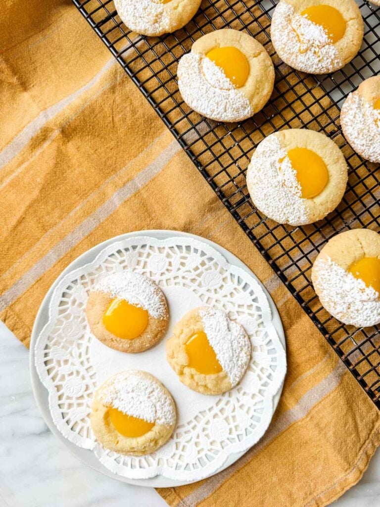 Lemon curd thumbprint cookies dusted with powdered sugar are sitting on a wire cooling rack and a small white plate with a round doily sheet.