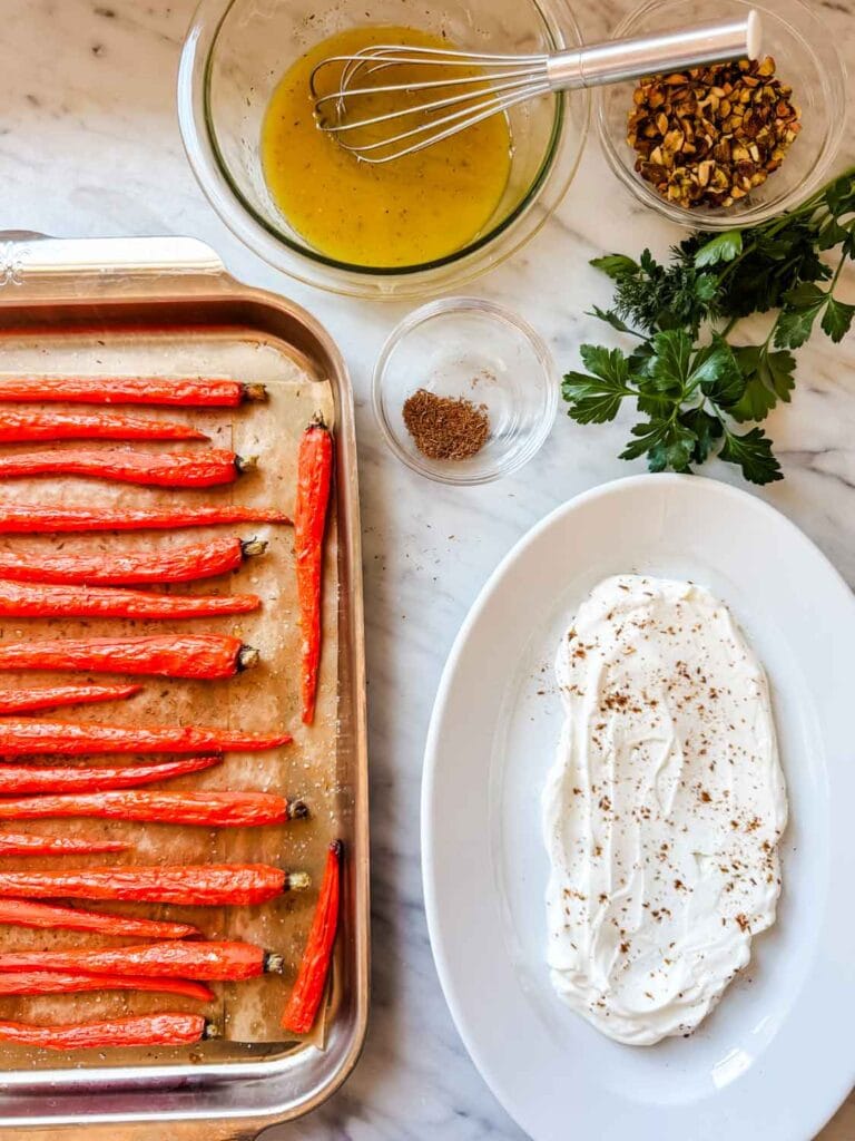 Roasted carrots on baking sheet, white platter with yogurt, bowl with dressing, herbs.