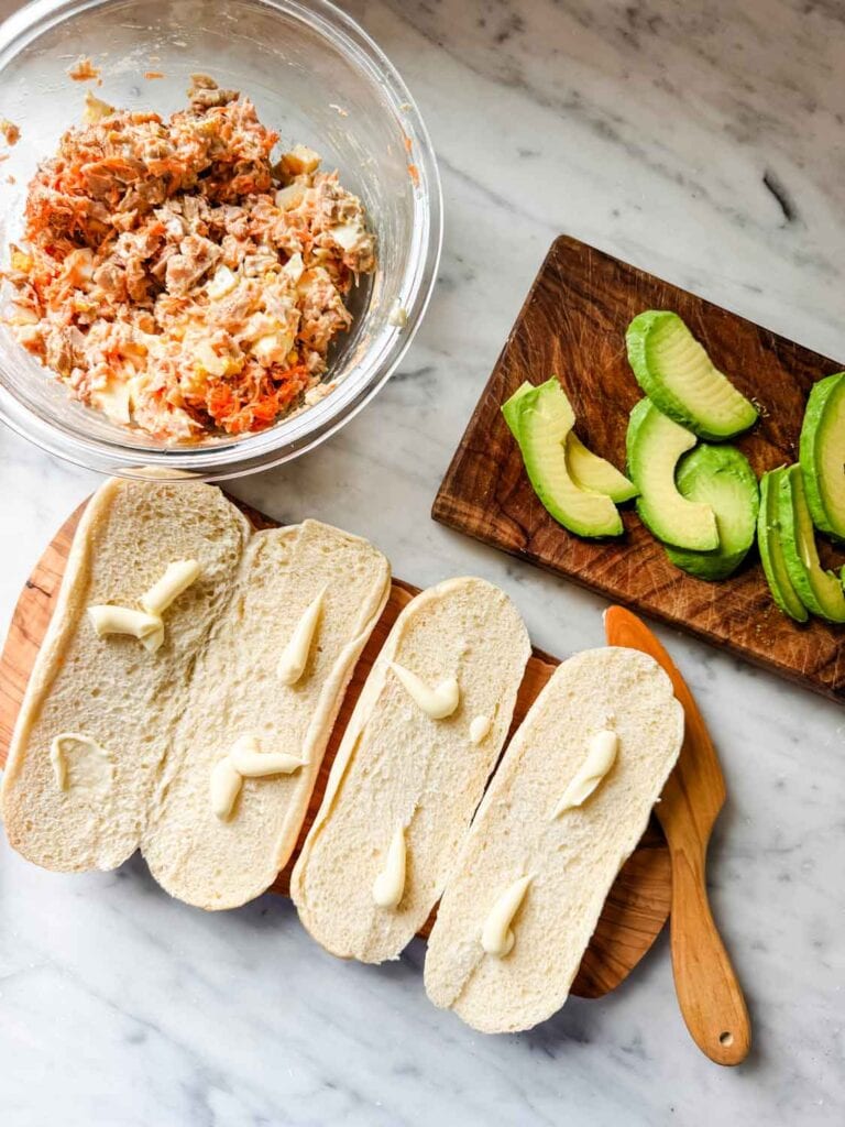 Bread rolls are open-faced and have a few small dollops of mayonnaise on them. Sliced avocado and a bowl of mixed tuna are sitting on the countertop. 