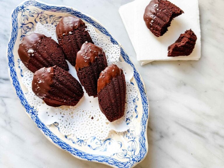 Double dark chocolate madeleines dipped in chocolate and sprinkled with sea salt are served on a vintage blue and white French plate. One Madeleine, broken in half, is sitting on a white napkin.