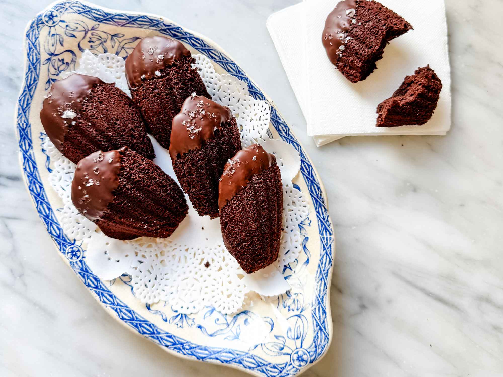 Double dark chocolate madeleines dipped in chocolate and sprinkled with sea salt are served on a vintage blue and white French plate. One Madeleine, broken in half, is sitting on a white napkin.