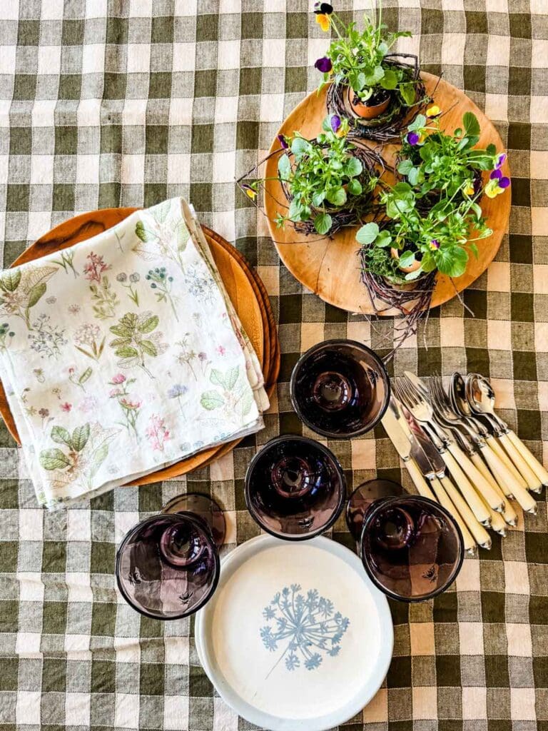 Napkins on wood plates, purple glasses, plants, cutlery on green and cream colred checked tablecloth.