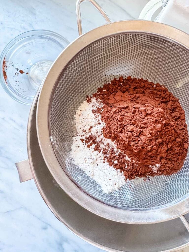 Flour, cocoa powder, salt, and baking powder are being sifted through a mesh strainer and into the the egg and sugar mixture.
