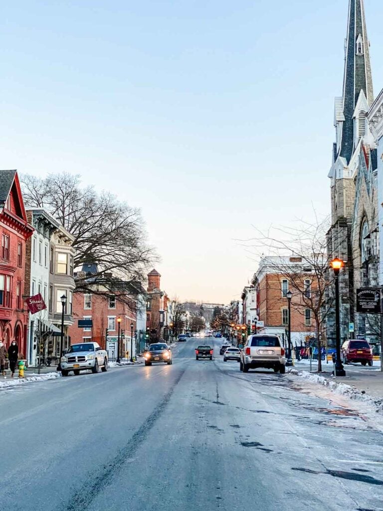 Main street in Hudson, New York in the winter with a car coming toward with headlights, streetlamp lit at dusk, some snow in the gutters.