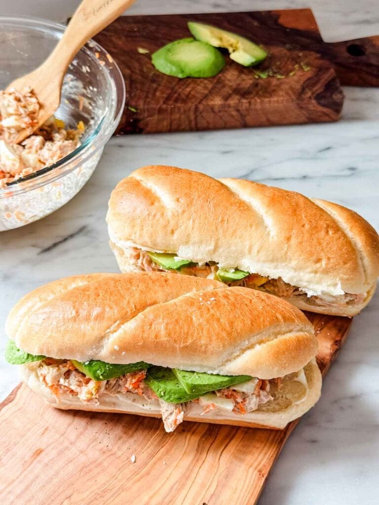 California tuna sandwiches are resting on a wooden cutting board. A bowl of tuna sits next to them on the kitchen counter. 