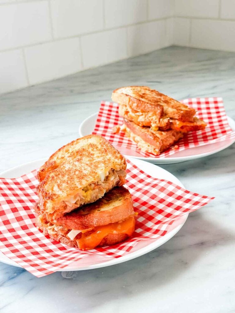 Two tuna melts are sliced in half, stacked on top of each other, and served on small plates covered with red-checked parchment paper.