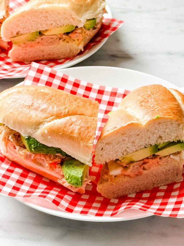 California tuna sandwiches are cut in half and served on small white plates with a sheet of red-checked parchment paper.