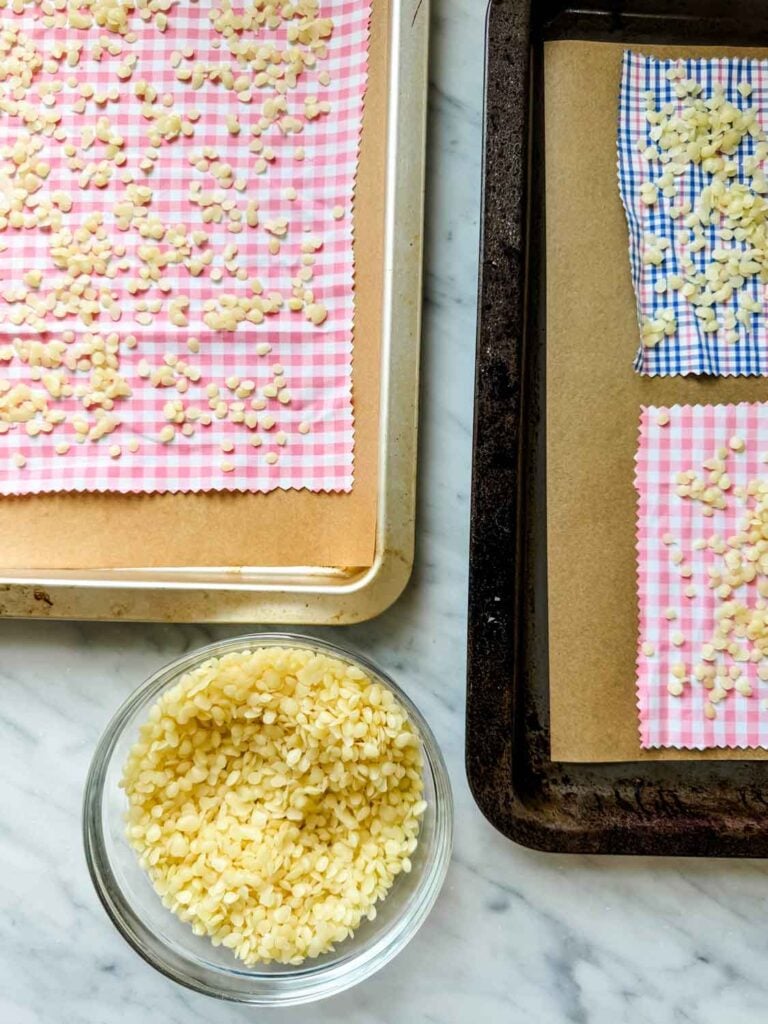 A bowl of beeswax pellets is sitting next to pieces of fabric with beeswax pellets evenly scattered on the fabric pieces.