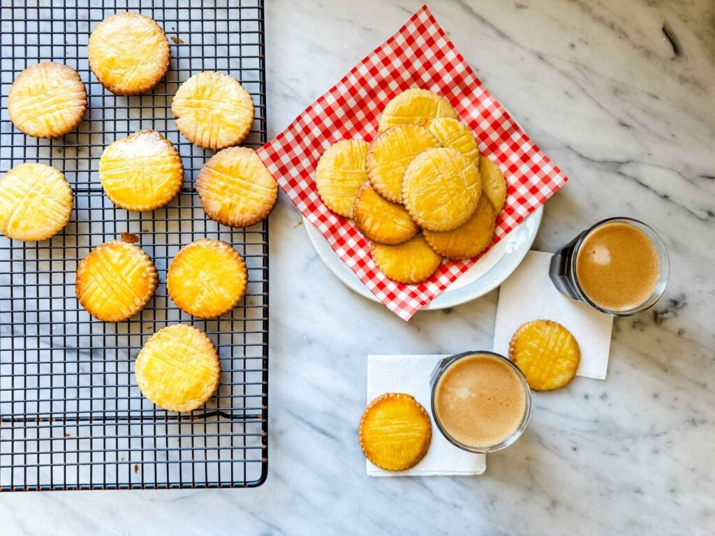 Freshly baked Sablés Breton cookies are on a red-checked piece of parchment paper on a vintage French plate. A cup of espresso is sitting next to the baked cookies.
