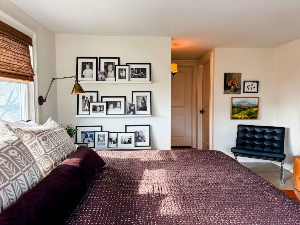 Bed, picture ledges wth black and white photos in frames, Knoll Barcelona chair. painting on wall, and a sconce.