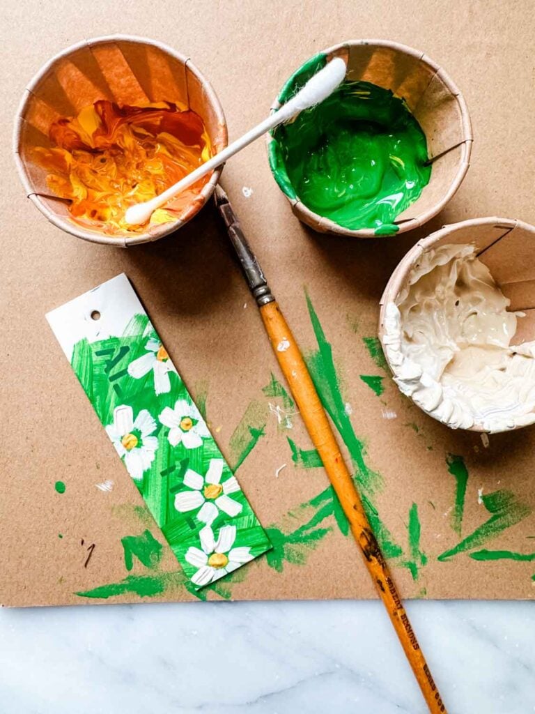 Yellow, green cream in cups with Q-tip resting on the paint cup. A tag with dasies and a paint brish on a background with green paint mess.