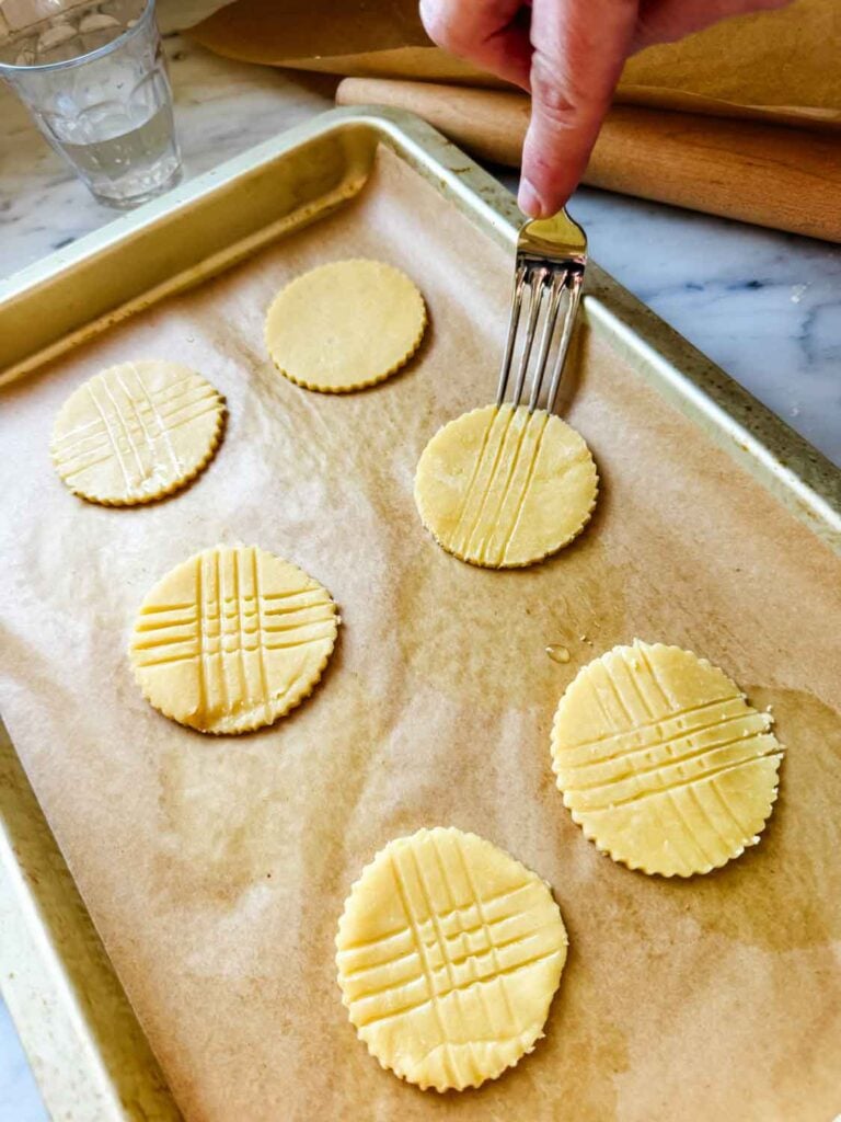 Fork tines, dipped in water, are used to create a pattern in the cut cookie discs that are on the parchment paper-lined baking sheet.