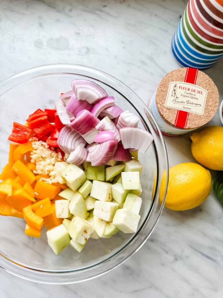 Chopped bell peppers, onions, garlic, and eggplant are in a glass bowl, ready for tossing with olive oil and roasting.