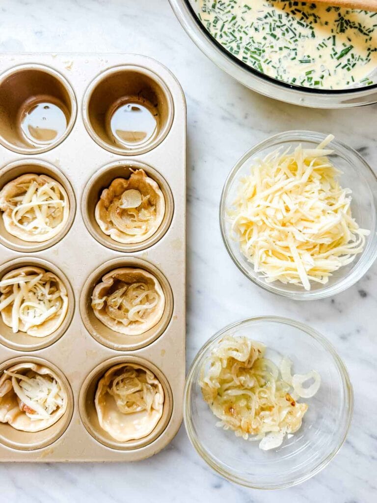Quiche in cupcake tins with small glass bowls of shredded cheese and vidalia onions.