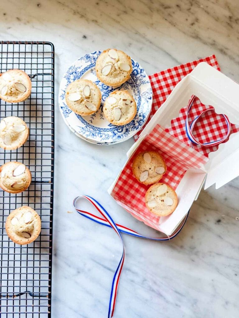 Baked almond tea cakes are on a cooling rack, a vintage French plate, and being packaged in a small, white pastry box.