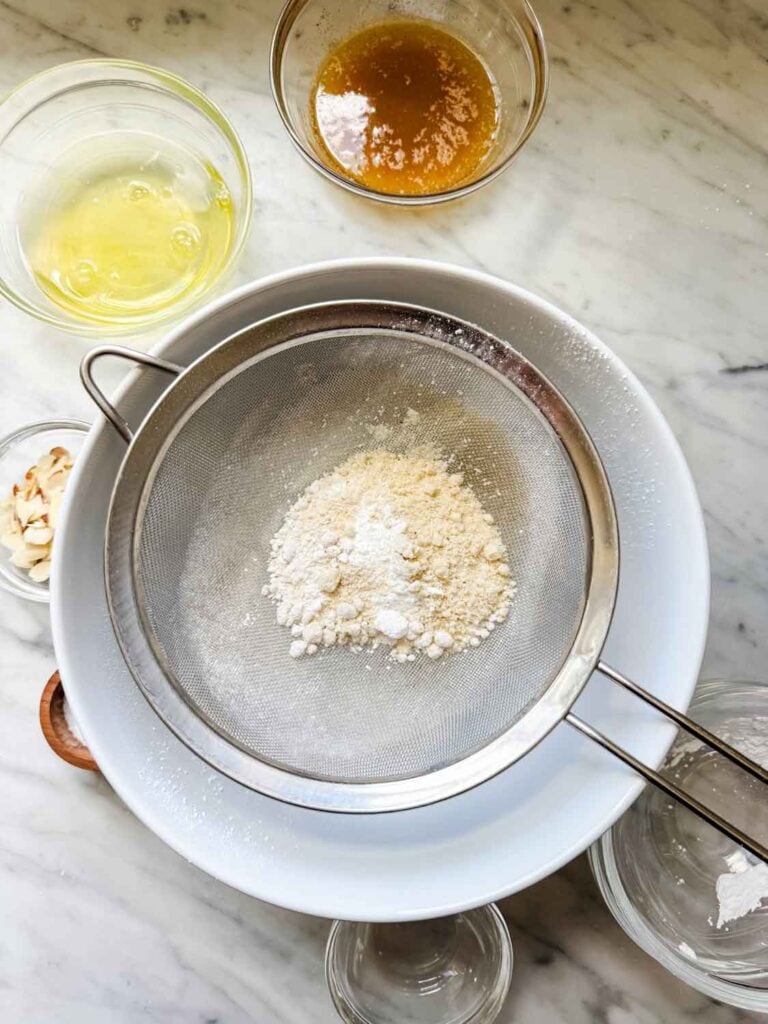 Powdered sugar, almond flour, regular flour, and salt are being sifted into a large, white mixing bowl.