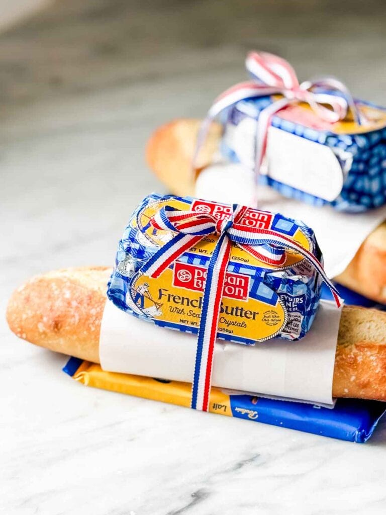 A fresh baguette is wrapped with a French chocolate bar and some good French butter. French red, white, and blue ribbon holds it all together for a small gift.