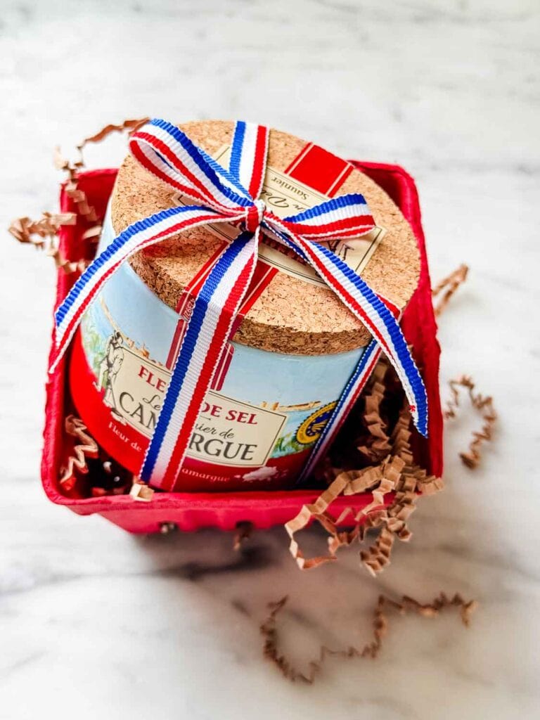 A small container of sea salt with a corked top is wrapped in a small cardboard crate with shredded paper and tied with a red, white, and blue French ribbon.