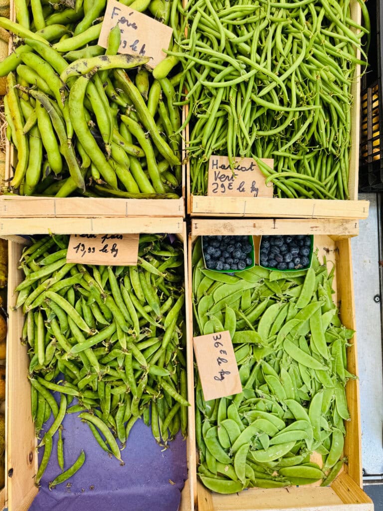 Green beans, haricots verts, and peas in their shells are in small wooden crates at Marché Raspail in Paris.