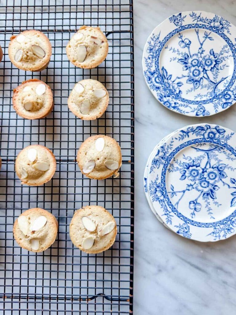 Classic French financier cakes are fresh from the oven and cooling on a wire rack next to vintage French plates.