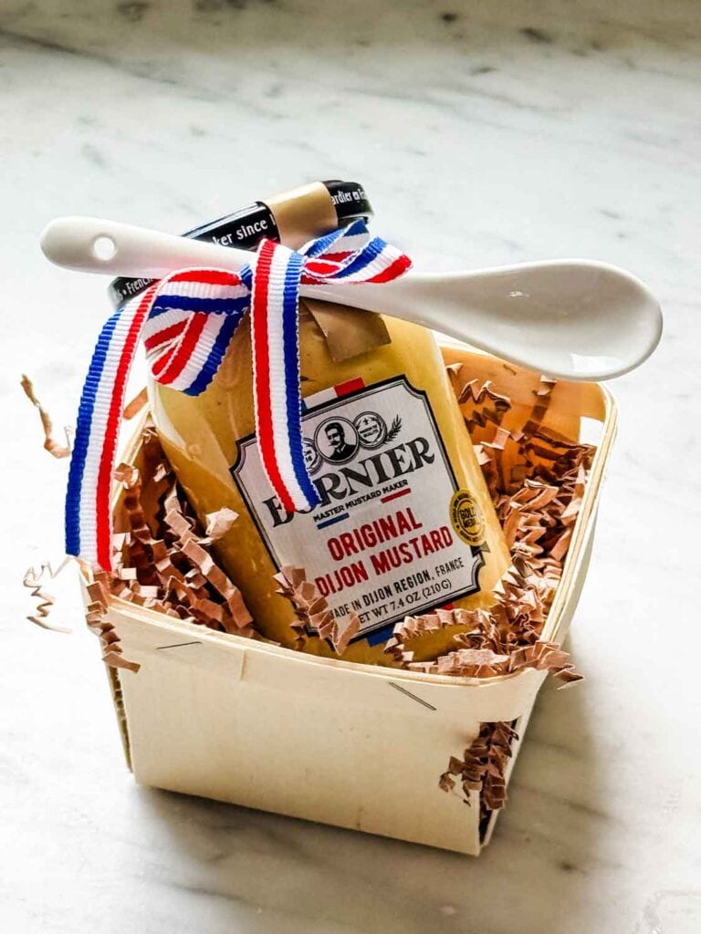 A jar of mustard with a small porcelain serving spoon is wrapped in a small wood crate with French ribbon for gifting.