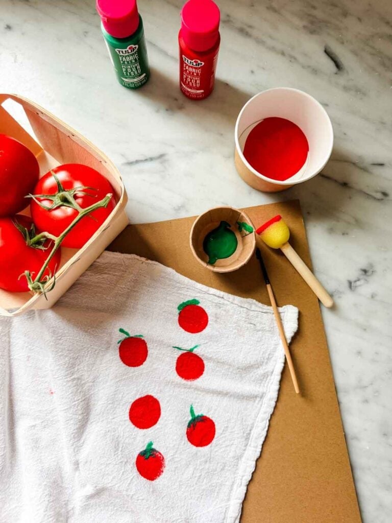 Tomatoes in a wooden berry crate, a whote towel with painted tomatoes, cups of red and green paint with a foam dauber and a paint brush.