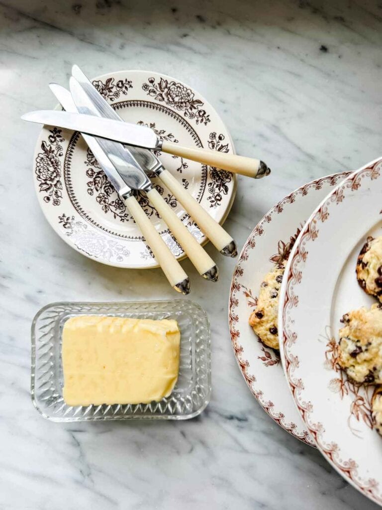 Small brown floral plates with cream handles knives resting on top, butter in glass dish, plates of pastry barely showing on the right side. 