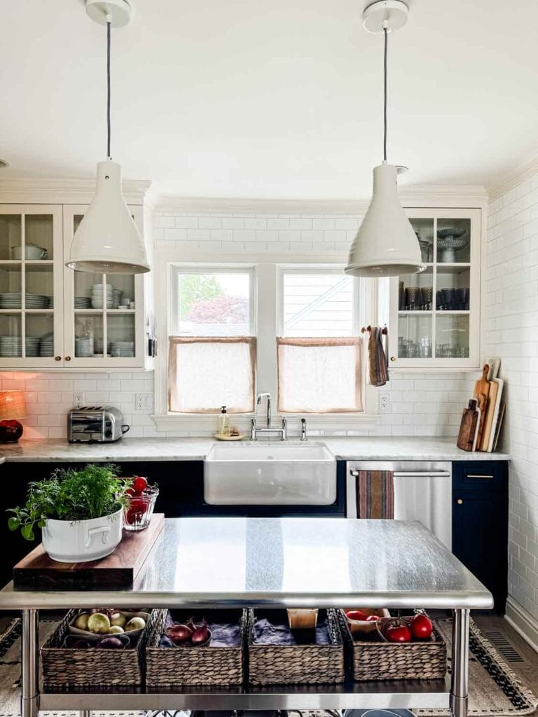 Stainless steel island in kitchen with farm sink, cafe curtains, ceramic pendants. 