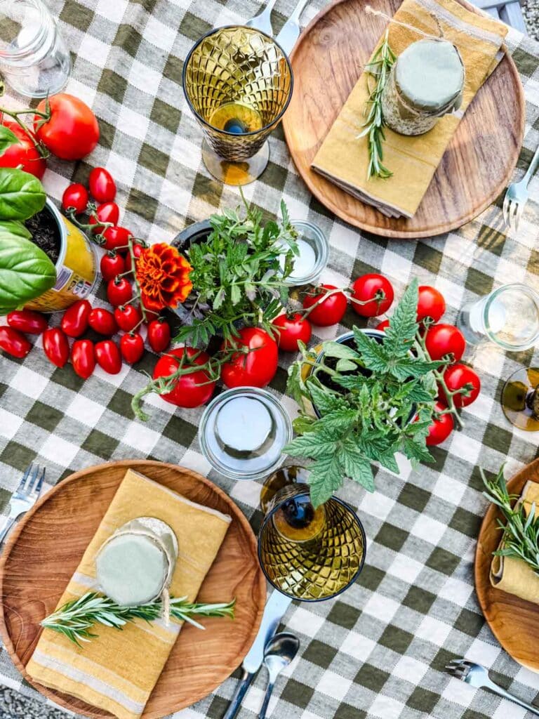 Small jar with votive, plants, tomatoes  on green checked tablecloth with wooden plates, gold linen napkins, rosemary and small jars.