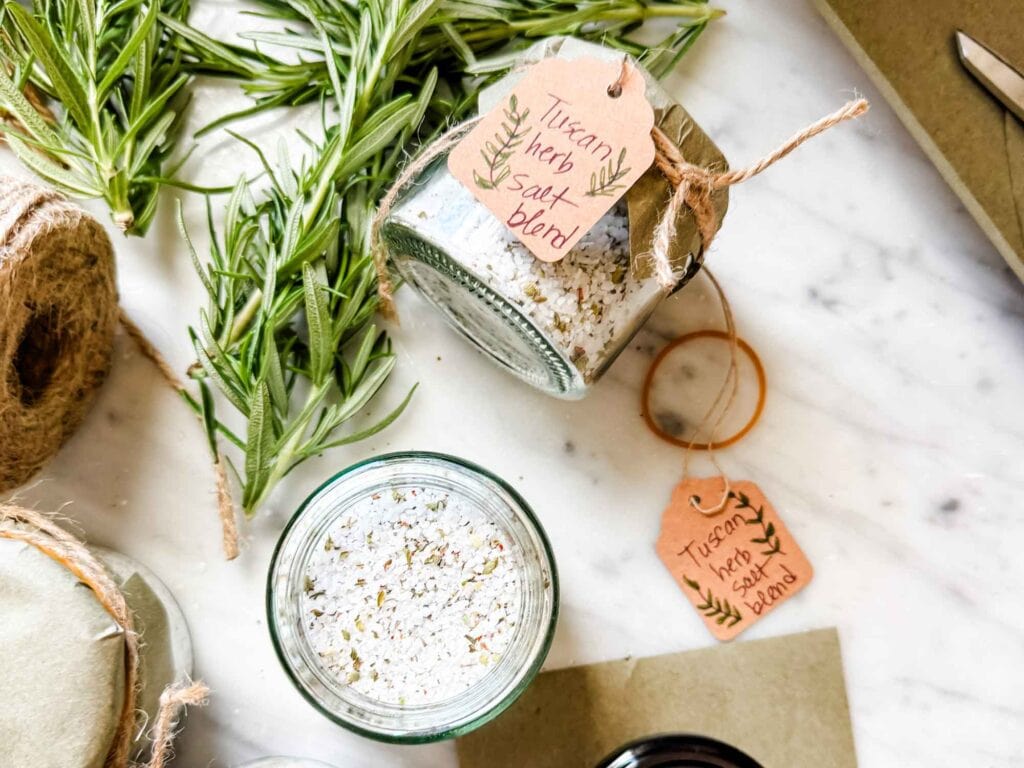 Jars of salt with small kraft colored tags, fresh rosemary, black lid and a rubberband on a marble counter.