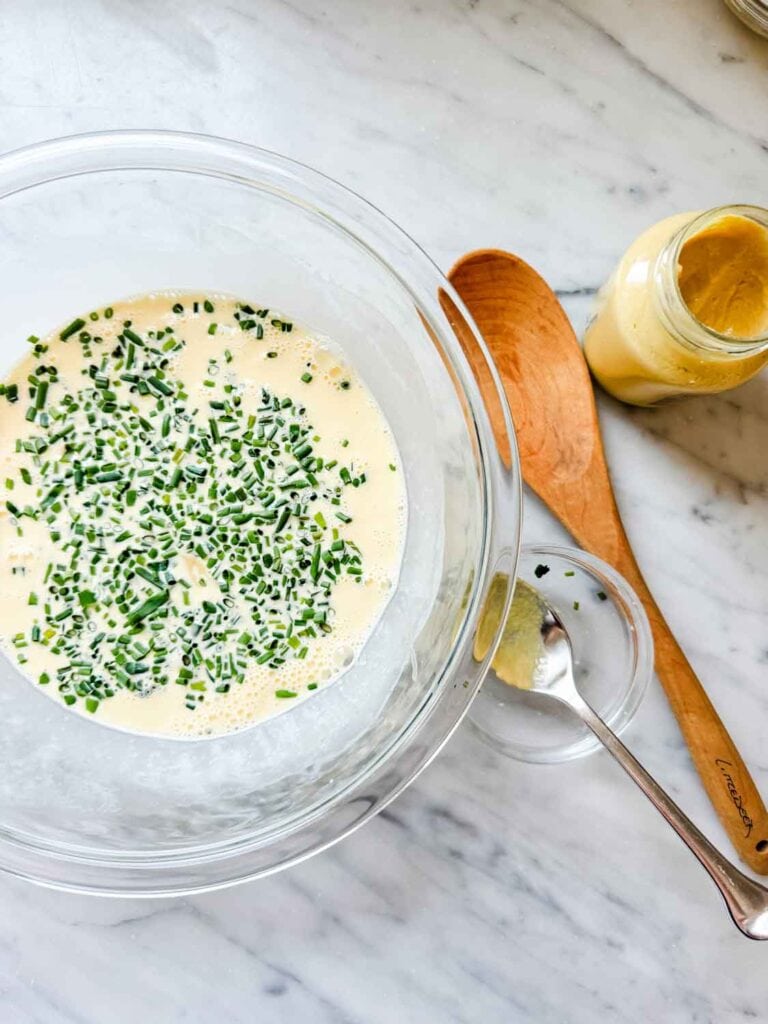 Mixing bowl with quiche filling with chives, mustard jar, spoon in small dish, plus a wooden spoon.